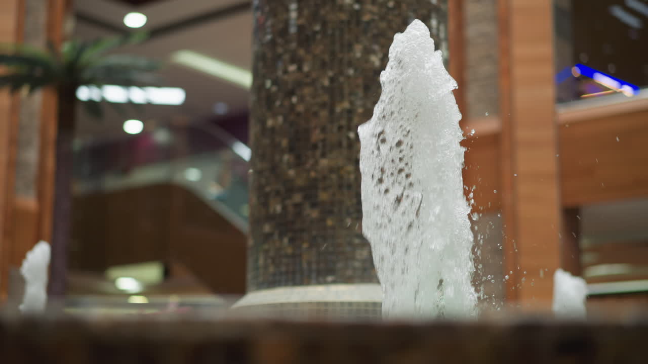 타일로 된 기둥과 현대적인 쇼핑몰 건축물로 둘러싸인 역동적인 물줄기를 가진 실내 분수, 흐릿한 초록색, 에스컬레이터 및 환경 조명