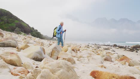 Mujer-Excursionista-Mayor-Con-Mochila-Y-Bastones-De-Senderismo-Caminando-Mientras-Camina-Por-La-Playa.