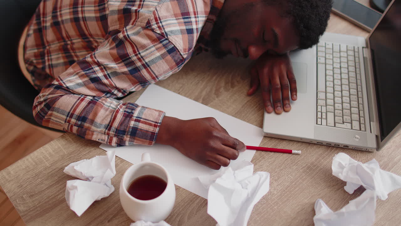 Tired man at home office falling asleep on table with laptop computer, crumpled sheets of paper