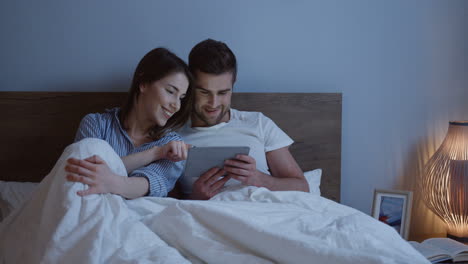 joven pareja caucásica sentada en la cama y viendo un video en la tableta por la noche