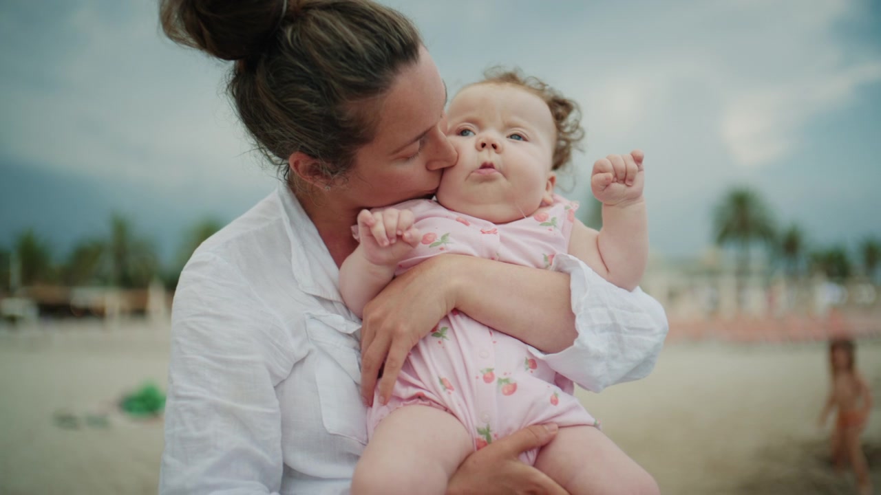 Bebê curioso inclinando-se para a frente enquanto é segurado por uma mãe perto da orla, observando as ondas suaves do oceano durante umas férias de praia em família