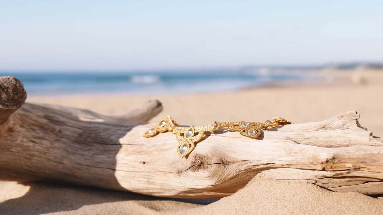 Kamera bergerak mundur menjaga kalung emas tetap fokus pada batang kayu apung yang memutih, menunjukkan pantai berpasir.