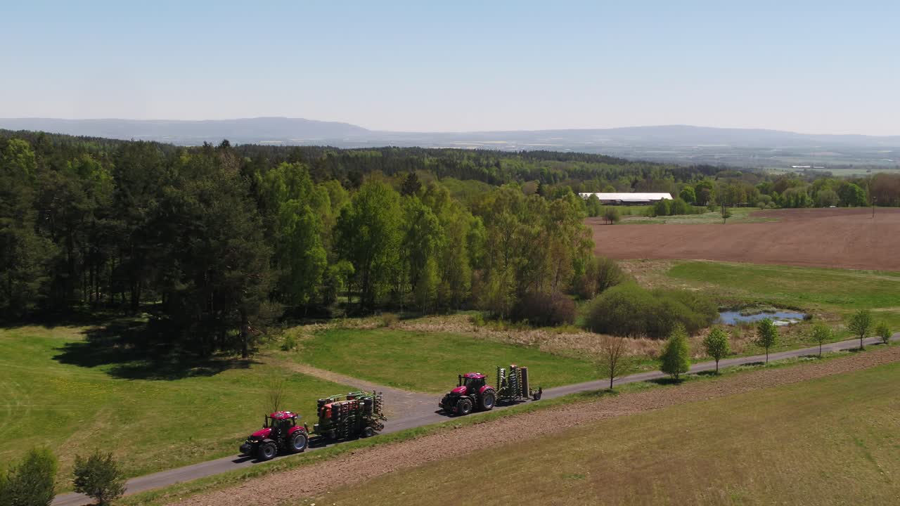 Drone zoom out of red tractors transporting folded agricultural equipment surrounded by green and brown fields