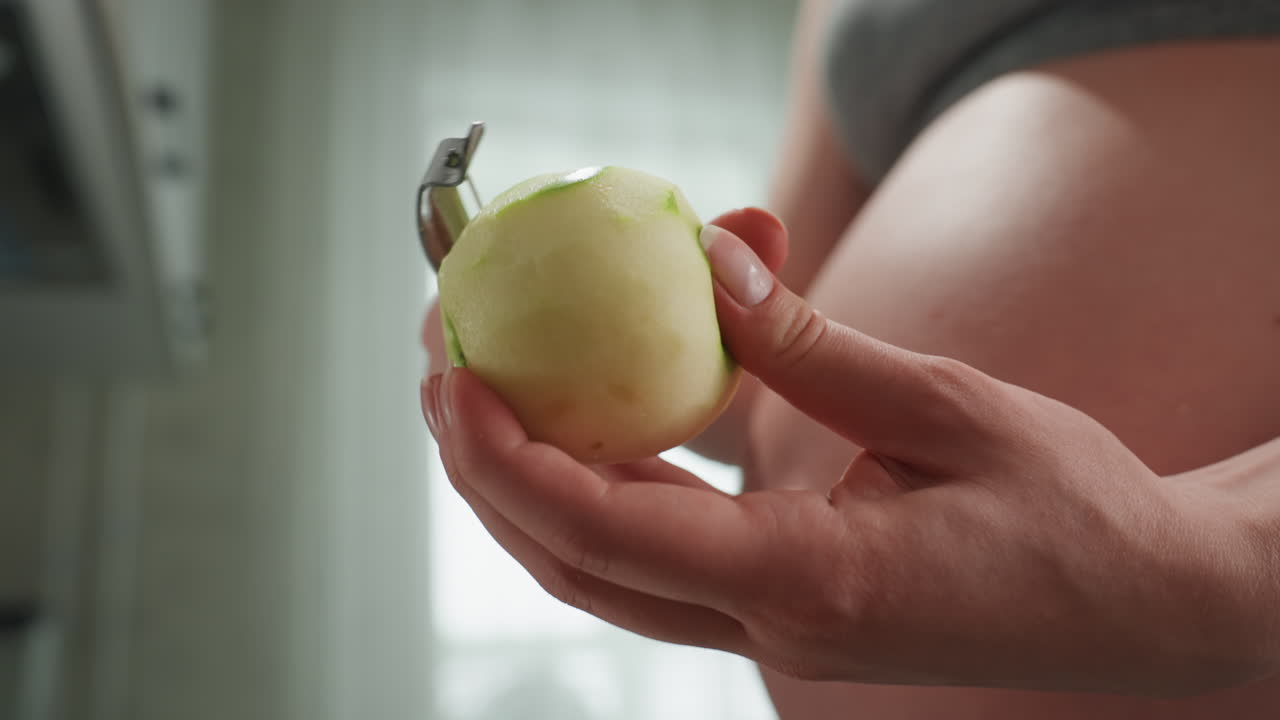 Primo piano sulla pancia bianca di una donna incinta che sbuccia una mela con mani attente e un pelapatate vicino al pancione scoperto, luce soffusa dalla finestra, la buccia che cade, attenzione alla nutrizione prenatale, consistenza e presa, cucina consapevole