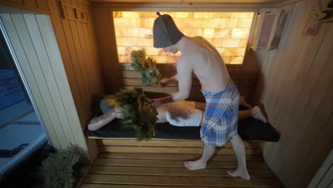 man lying on wooden bench in spa with masseur doing massage with oak broom. client in finn sauna indoors with professional.