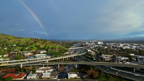 Regenbogen-Skyline-über-Der-Autobahnbrückenkreuzung-In-Zeitlupe,-Stadtverkehr,-Stadt-Mit-Klarer-Skyline,-Aufnahme-Aus-Der-Luft-Mit-Einer-Drohne