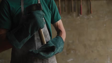 midsection of caucasian male blacksmith holding hammer in workshop