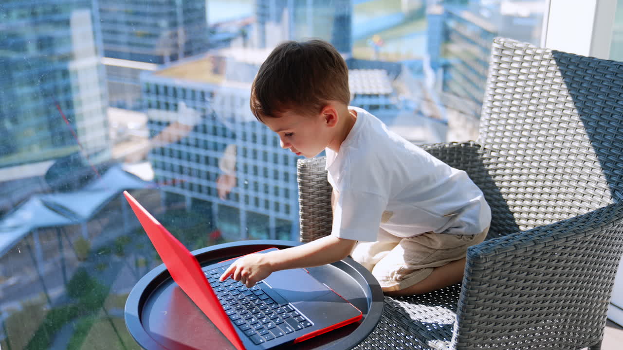 Dark-haired Caucasian baby sits on his knees in the armchair. Cute kid looks at laptop and then explores keyboard