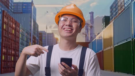primer plano de un trabajador asiático con gafas de protección y casco de seguridad sonriendo y señalando un teléfono inteligente en la mano mientras está de pie en el almacén del patio de contenedores
