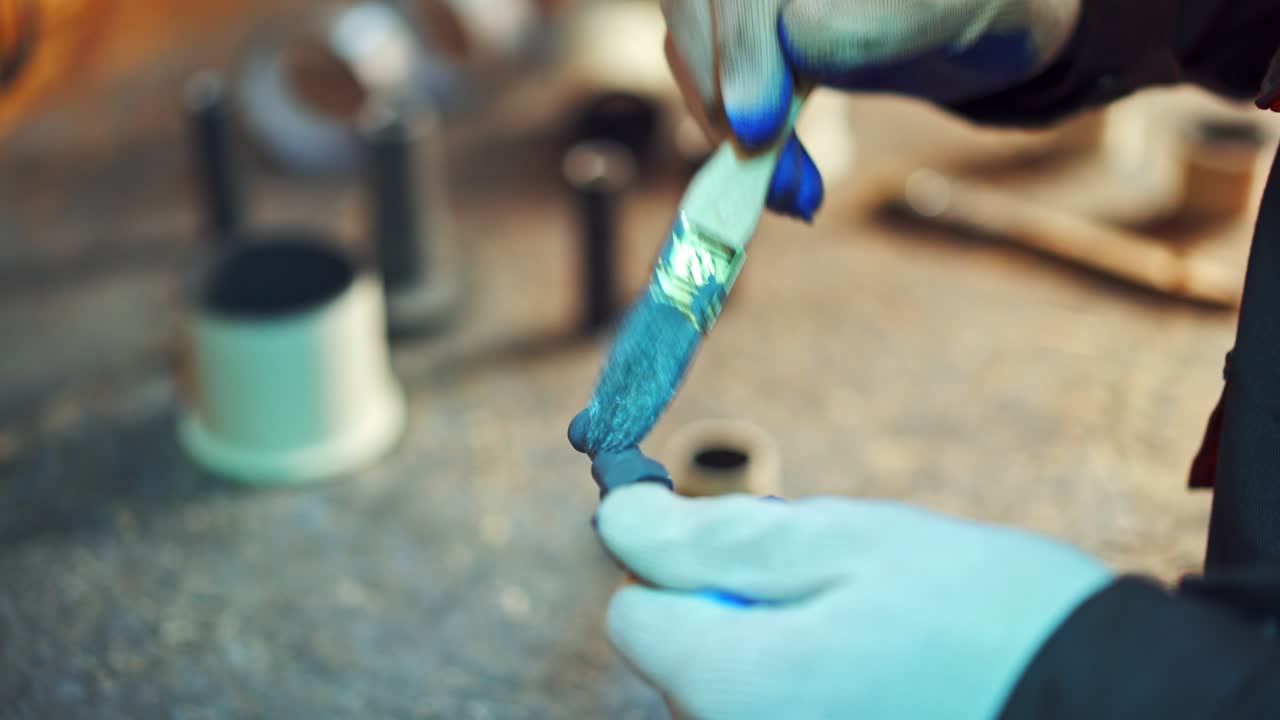 Close-up of man's hands with a brush painting metal tool with blue color in factory. Male worker in gloves paints instruments at workshop.