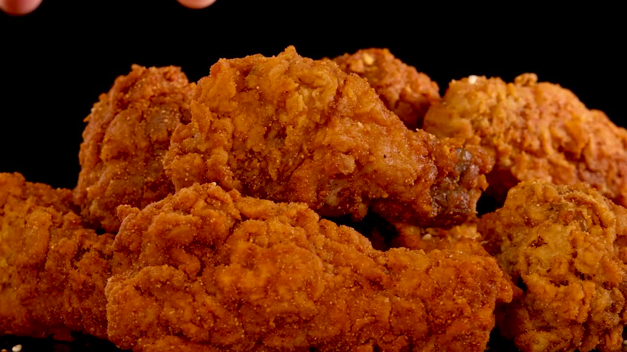A hand reaches in to pick up a crispy, spicy fried chicken piece from a pile under dramatic studio lighting, with a black background and slow motion effect