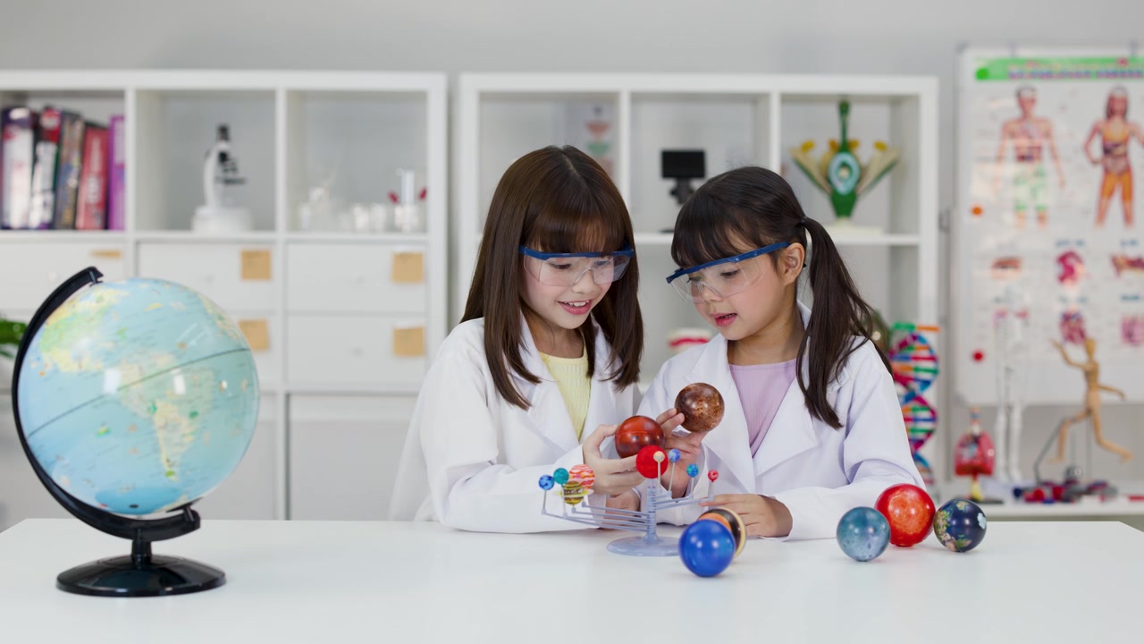 Dua gadis muda mengenakan jas lab dan kacamata pelindung memeriksa model planet di ruang kelas