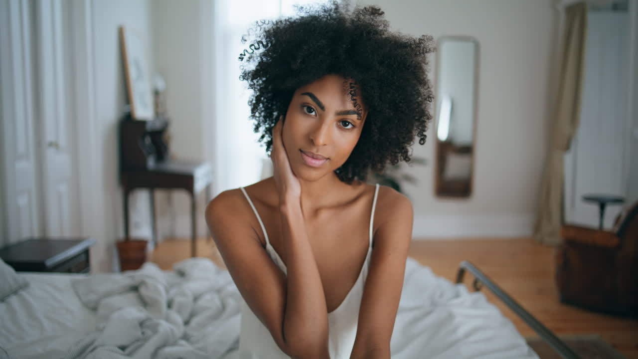 Modella delicata seduta a letto, ritratto casalingo. Donna africana che si tocca i capelli scuri e ricci.