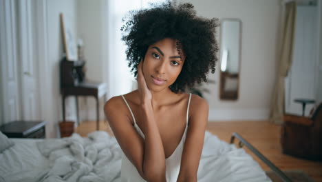 gentle model sitting bed home portrait. african woman touching dark curly hair