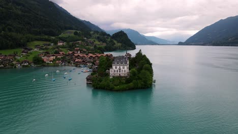 Castillo-De-Iseltwald-En-Una-Península-En-Un-Lago-Suizo,-Con-Un-Telón-De-Fondo-De-Un-Pintoresco-Pueblo-Y-Montañas,-Vista-Aérea