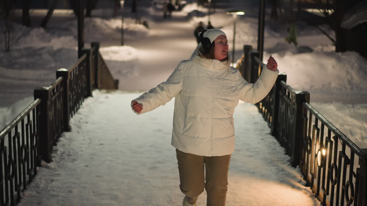 Cheerful woman in white puffer coat and headphones dancing on snow covered pavilion stairs at night with faint silhouettes in background moving through illuminated winter park scene gracefully
