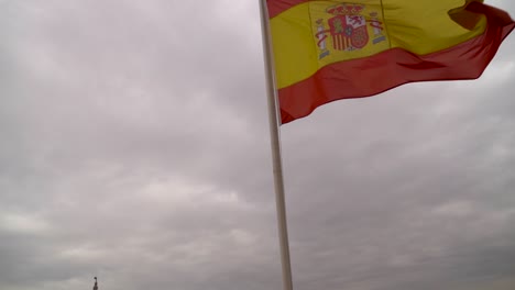 Inclinación-Lenta-Hacia-Abajo-Desde-La-Bandera-Española-Ondeando-Contra-El-Cielo-Gris-Con-Nubes-Y-Ciudad-En-La-Distancia