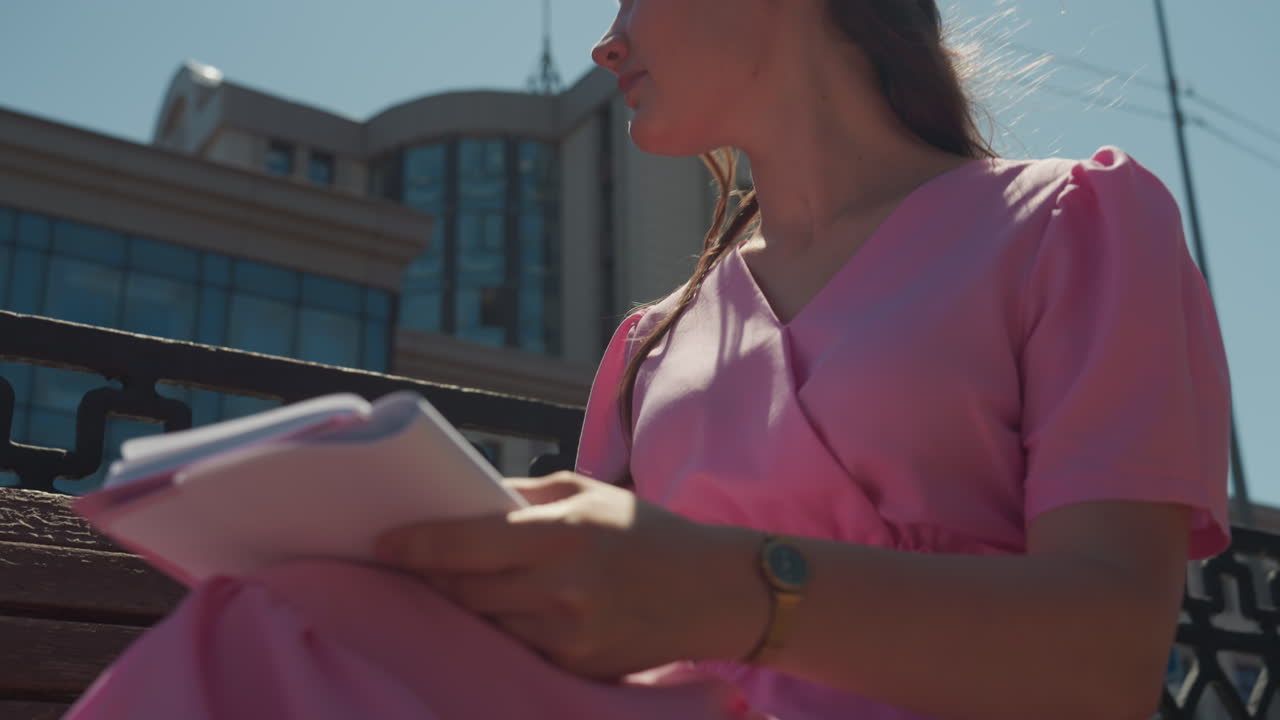 Mulher caucasiana escrevendo anotações em um banco da cidade, close-up das mãos e do lápis, caderno aberto no colo, vestido rosa, detalhe do relógio de pulso, luz solar forte de verão, postura concentrada, calçada e arquitetura urbana