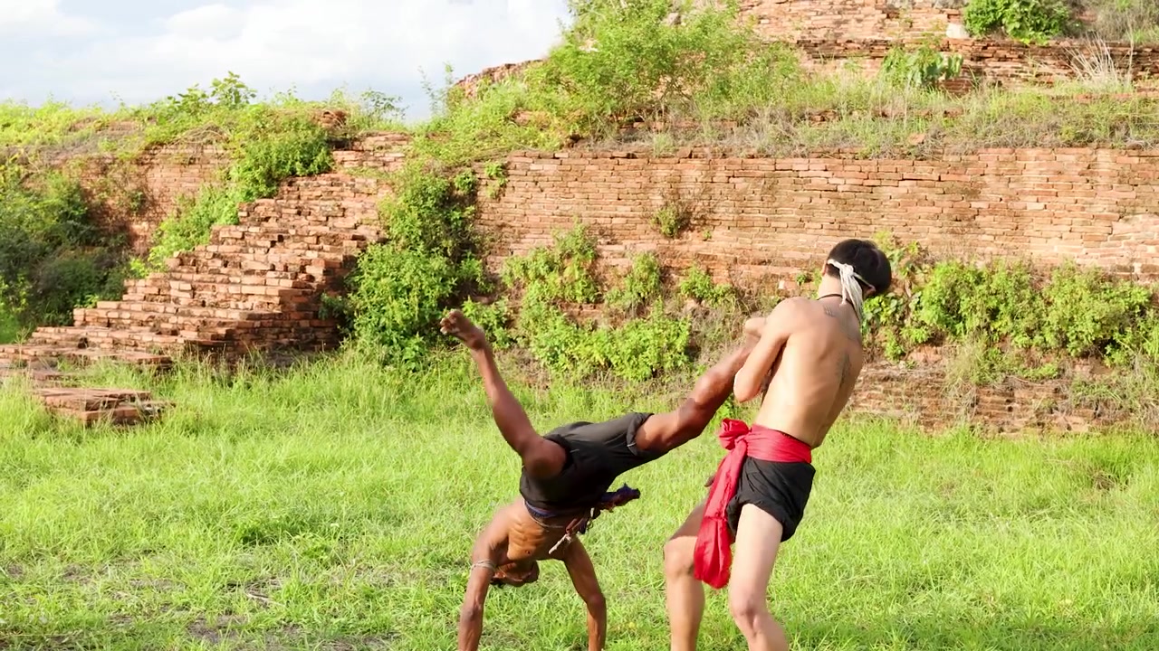 Two men practice Muay Boran techniques outdoors against a historic brick temple wall backdrop.
