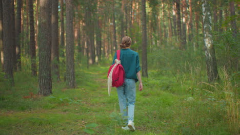 vista trasera de una mujer caminando por un tranquilo bosque iluminado por el sol, mochila roja colgada sobre el hombro, camisa verde mezclada con la naturaleza, la luz del sol se filtra a través de los árboles y la hierba alta