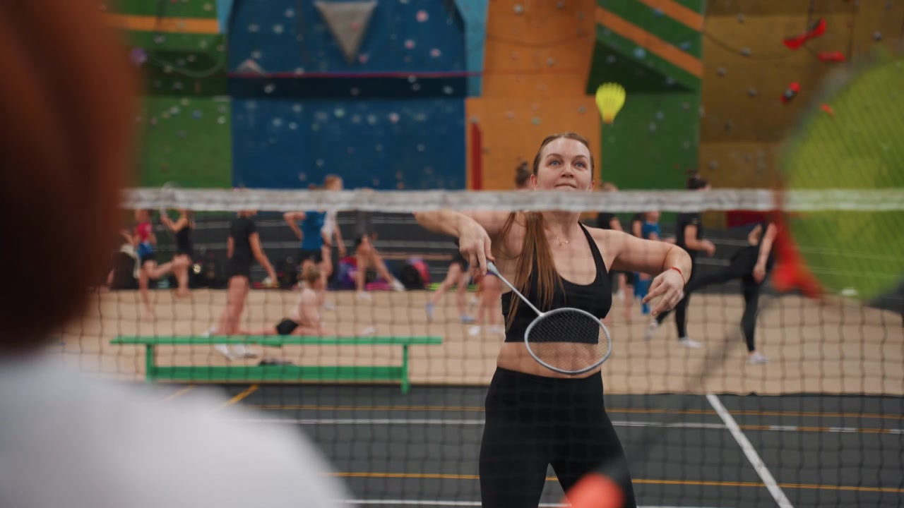 Indendørs badmintonnet, intens kvindelig atlet, nærbillede af volleyudvekslinger, kraftfulde sving og fokuseret udtryk, tilskuere i gymnastiksalen og levende vægpaneler, højenergisk konkurrence.