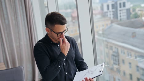 young dark-haired man in black shirt holding papers in his hands. male employee touches glasses and looks at the window. high angle view.