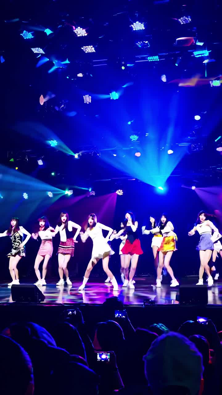 Kpop girl group captivating fans while dancing on stage during a vibrant concert, surrounded by colorful spotlights and enthusiastic audience members recording the performance with smartphones