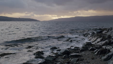 Orilla-Del-Agua-De-Mano-Y-Rocas-Al-Atardecer-Con-Tierra-Distante