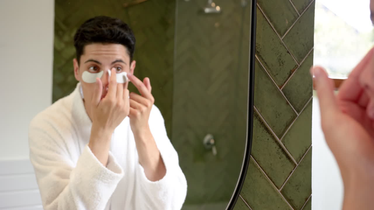 Focused biracial man applying under eye masks looking in bathroom mirror, slow motion