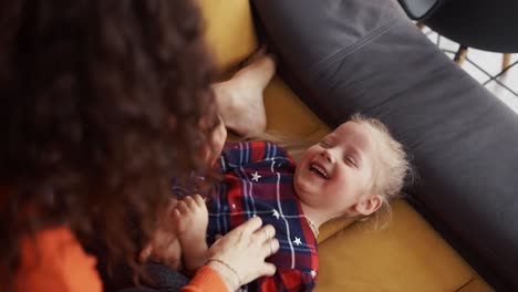 loving mother tickling baby daughter little girl on sofa, sincerely laughing
