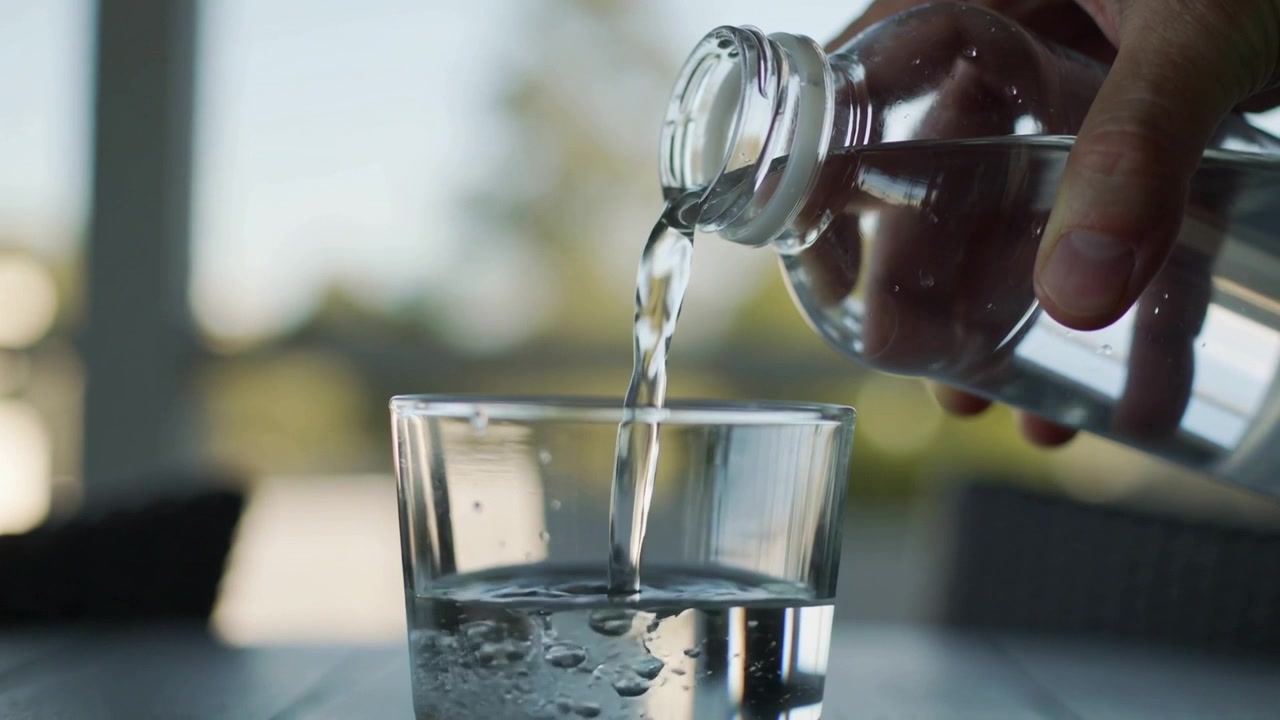 Eine rechte Hand neigt eine Flasche und gießt Wasser in ein Glas auf einem Tisch nahe dem Fenster, wodurch das Glas gefüllt wird.