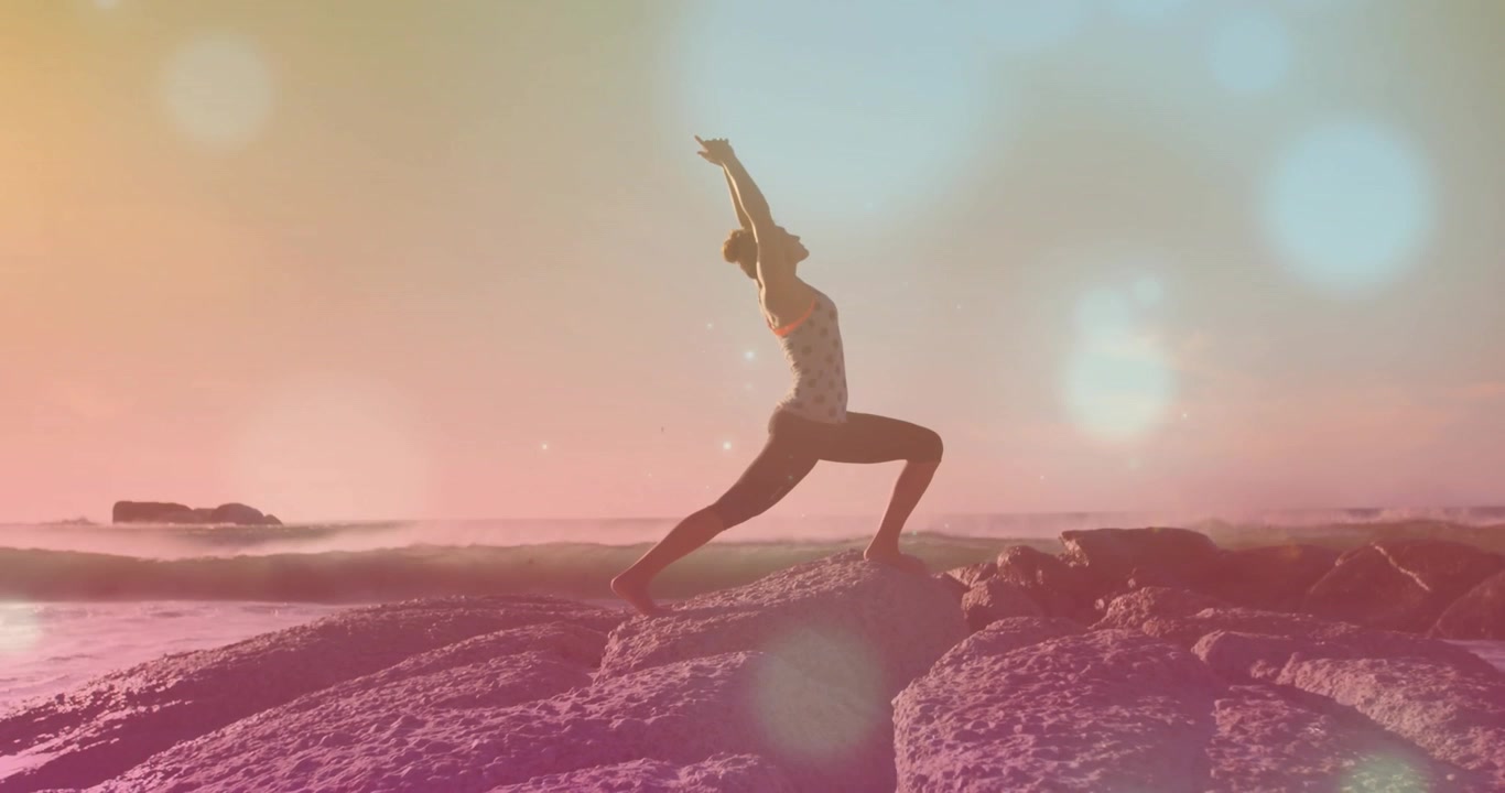 Donna che fa yoga in affondo alto su una riva rocciosa, con la luce del sole che crea un bagliore per l'equilibrio