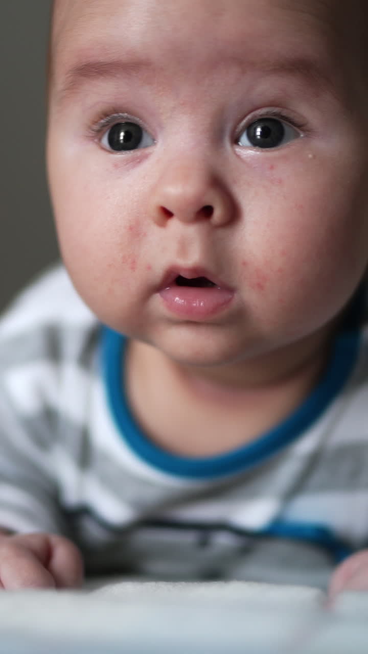 Interested look of a little toddler lying on his belly. Curious boy tries to keep his head and stares up. Close up. Vertical video
