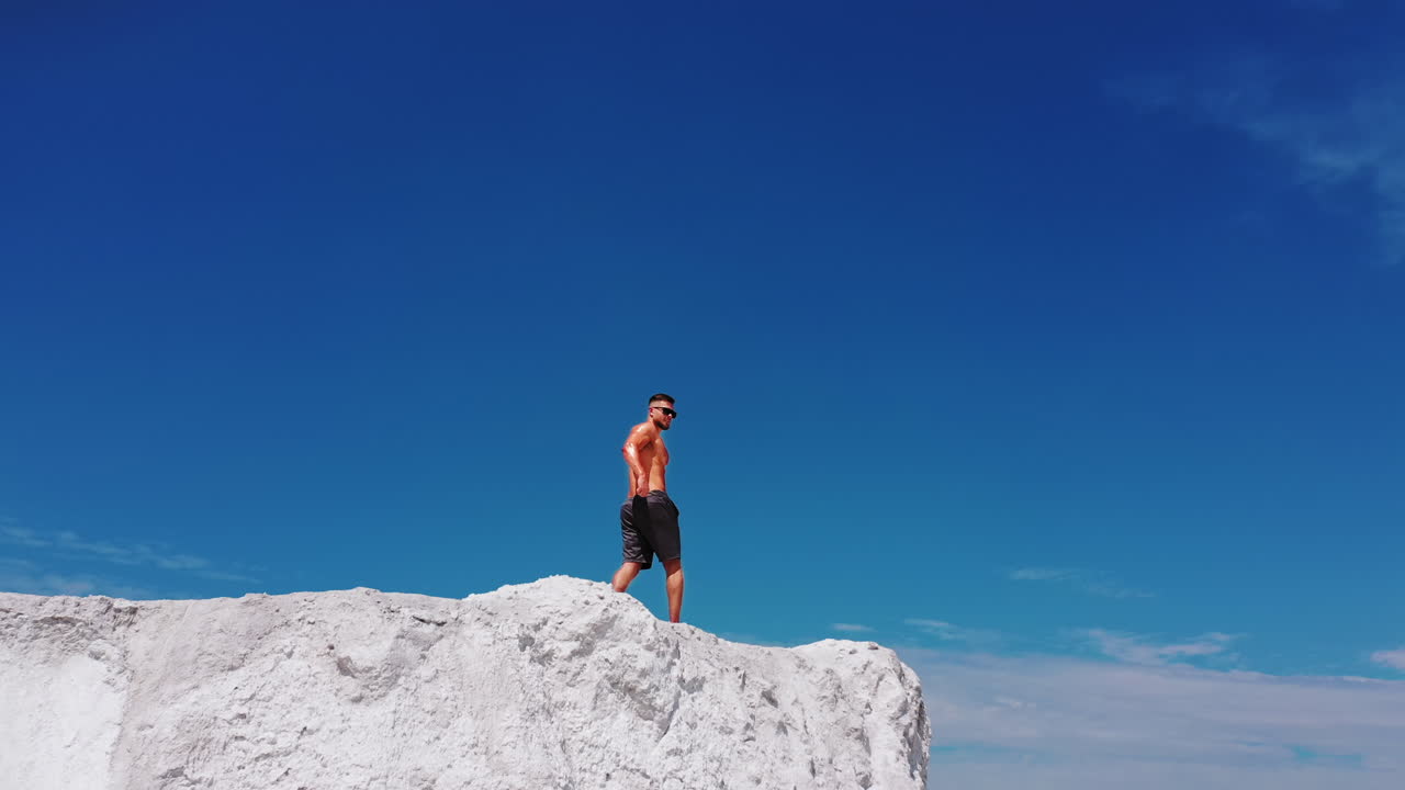 Muscular man on white hill. Bodybuilder without shirt posing on the top of the mountain under blue sky. Motion camera back.