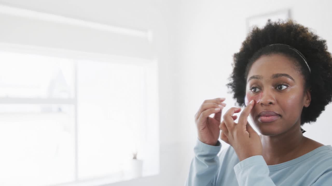 Happy african american woman applying under eye mask looking in bathroom mirror, slow motion
