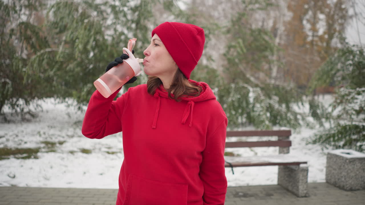dama con capucha roja y gorra beba agua de botella rosa al aire libre en el parque de invierno con suelo nevado, árboles verdes y banco en el fondo