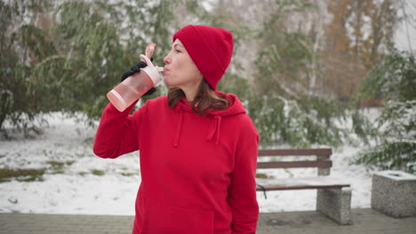 lady in red hoodie and beanie sips water from pink bottle outdoors in winter park setting with snowy ground, green trees, and bench in background