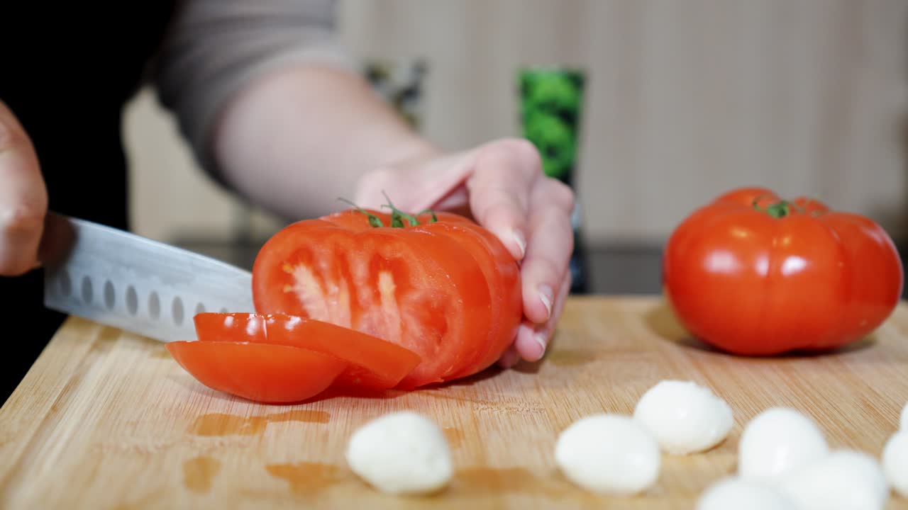 Hand slices ripe tomato with sharp knife next to peeled onions on cutting board