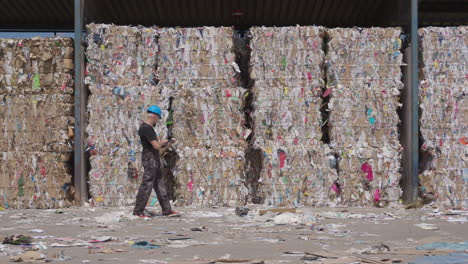 Worker-walks-by-stacked-paper-bales-at-recycling-plant,-wide-slomo