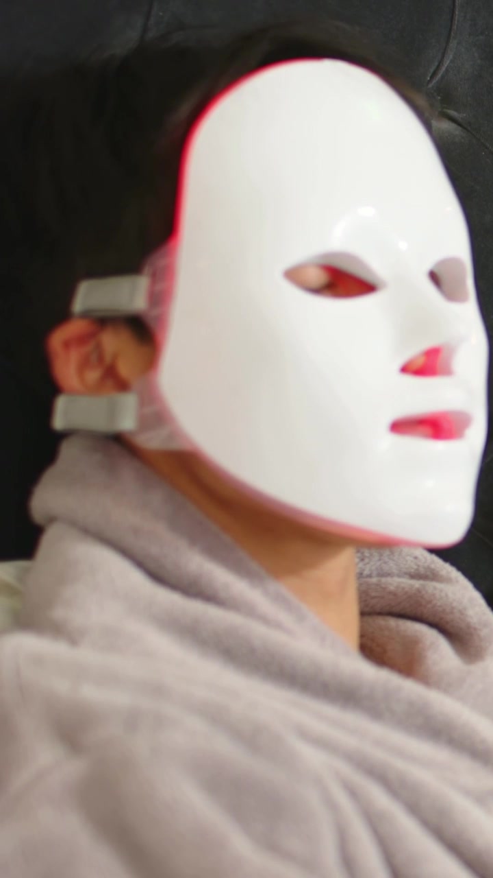 Vertikales Video: Liegender Mann trägt LED-Maske im Schlafzimmer, erhält Rotlichttherapie und blinzelt
