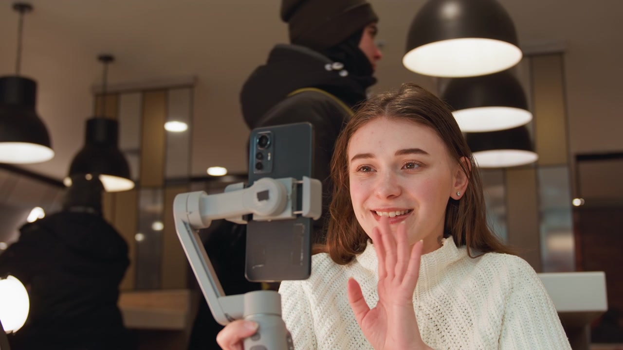 Mujer sonriente sostiene un smartphone en un estabilizador de mano y saluda hacia el dispositivo durante una creación amigable dentro de un cálido espacio de trabajo interior, mostrando un agarre firme, estilo casual, luz suave y compromiso relajado.