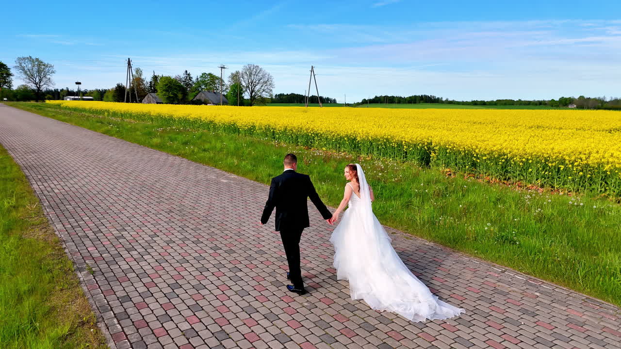 Sarı çiçek açmış bir tarlanın yanındaki Arnavut kaldırımlı bir yol boyunca el ele yürüyen bir gelin ve damadı takip eden yer seviyesinden bir çekim, bahar sıcaklığını, açıklığı ve umut dolu, romantik bir dinginliği yakalıyor.