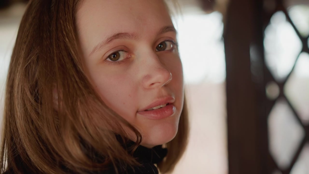 Near-frontal portrait of young woman turning toward camera while loose brown hair frames face, soft gaze and faint smile creating calm confident mood in warm indoor window light, with woman, portrait