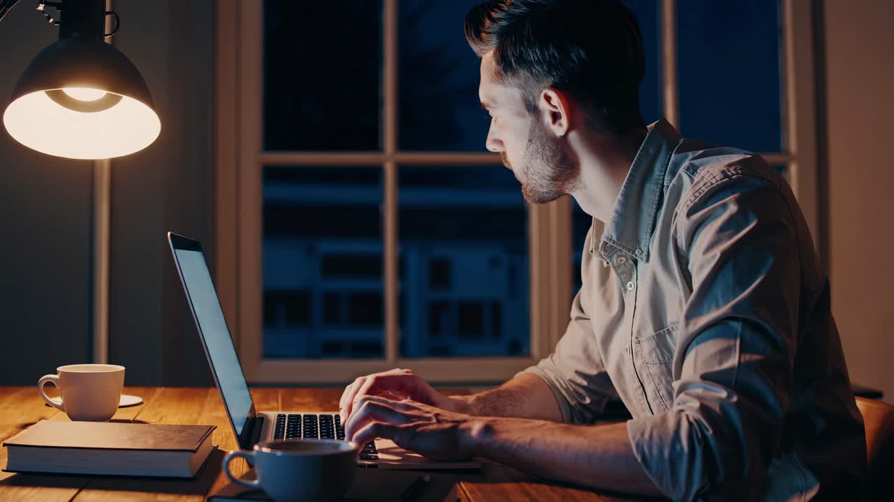 A man works intently on a laptop at night, illuminated by a desk lamp