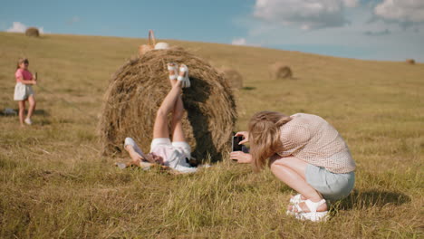 rear view of woman taking photo of friend lying on hay bale with legs up, picnic basket and hat beside them, a little girl holding a dog leash stands nearby in golden field under blue sky