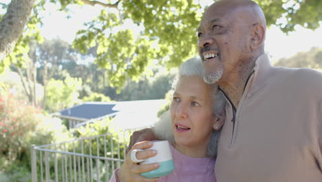 happy senior biracial couple embracing and drinking coffee at balcony at home, slow motion