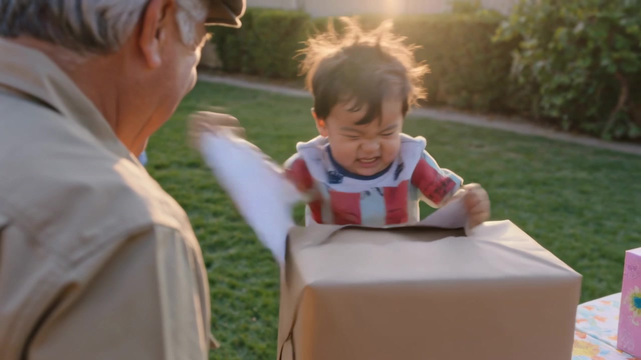 Bambino con bavaglino e maglietta a righe che tocca una scatola incartata, strappando la carta velina bianca e trovando un regalo a un tavolo in giardino.