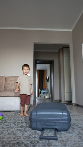 cute toddler boy walking around the suitcase in the room. little kid sits down to play with valise wheel. vertical video.