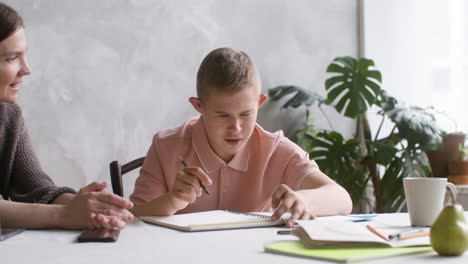 close-up view of a boy with down syndrome doing homeworks sitting at table in the living room at home. his mother helps him