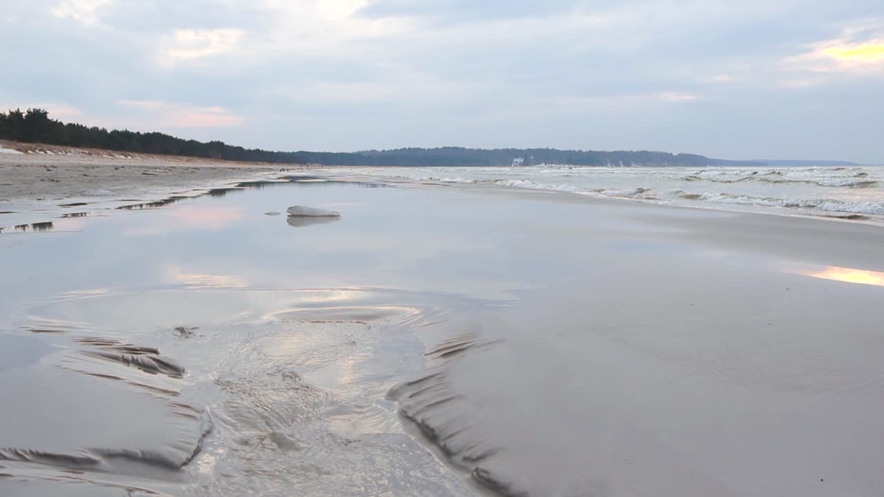 Gorgeous low angle footage of a sandy beach shoreline in Vääna jõesuu during cloudy autumn or spring day with water flowing and reflecting on the sand, while Baltic sea ocean waves are moving to shore